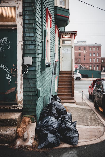A small urban residential building with a teal-colored wooden exterior featuring horizontal siding and a white front door accessed via a short staircase with wooden steps. To the left of the entrance, a small gray utility box is mounted on the wall, along with some cables and a black electrical box beneath it. Several black garbage bags, filled and tied, are piled on the sidewalk at the base of the building next to a concrete step. The sidewalk area shows signs of dirt and a small patch of snow or ice near the bags. In the background, a parking lot with a few parked vehicles, including a dark-colored car on the right, is visible, along with a brick industrial-style building further in the distance. Overhead, power lines streak across the sky, and the overall atmosphere appears overcast, typical of an urban environment where private waste collection or refuse removal services like those offered by rubbishremovalkingstonuponthames.org.uk may be employed as an alternative to local authority rubbish collection.
