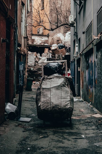 A narrow urban alleyway with dark, uneven pavement, lined by two buildings—one on the left with exposed brickwork and the other on the right with painted and graffiti-covered walls. In the foreground, there is a large, wheeled black and grey trash bag or bin, which appears full and slightly torn, with cables or straps visible on it. Behind this, cluttered piles of cardboard boxes, plastic bags, and miscellaneous waste materials are stacked against a makeshift barrier or wall at the back of the alley. The cardboard boxes vary in size and are partially flattened, some piled irregularly, with some showing signs of wear and dirt. The scene is outdoors, with a faint overcast sky visible above, and a few leafless tree branches extend into the frame from the background, indicating a winter or late autumn setting. The overall atmosphere is gritty and cluttered, reflecting a typical area where private waste collection or rubbish removal services might be required, such as for on-site clearance or alternative waste handling outside of municipal collection areas. The presence of graffiti suggests a neglected or less-frequented urban location, emphasizing the need for professional rubbish removal in the area.