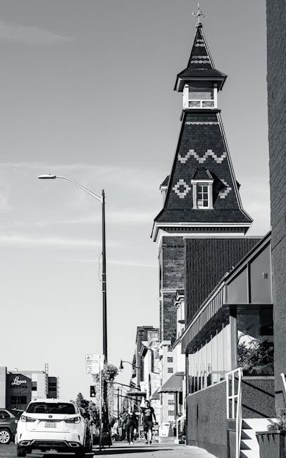 A narrow urban alleyway with dark, uneven pavement, lined by two buildings—one on the left with exposed brickwork and the other on the right with painted and graffiti-covered walls. In the foreground, there is a large, wheeled black and grey trash bag or bin, which appears full and slightly torn, with cables or straps visible on it. Behind this, cluttered piles of cardboard boxes, plastic bags, and miscellaneous waste materials are stacked against a makeshift barrier or wall at the back of the alley. The cardboard boxes vary in size and are partially flattened, some piled irregularly, with some showing signs of wear and dirt. The scene is outdoors, with a faint overcast sky visible above, and a few leafless tree branches extend into the frame from the background, indicating a winter or late autumn setting. The overall atmosphere is gritty and cluttered, reflecting a typical area where private waste collection or rubbish removal services might be required, such as for on-site clearance or alternative waste handling outside of municipal collection areas. The presence of graffiti suggests a neglected or less-frequented urban location, emphasizing the need for professional rubbish removal in the area.