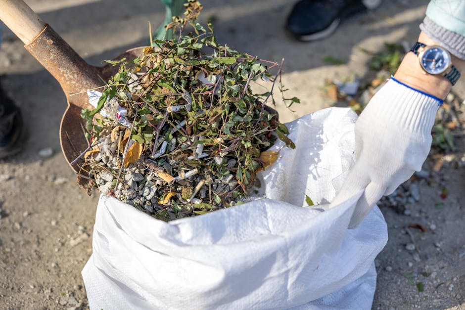 A person wearing a white glove is holding a large white sack filled with garden debris, including small branches, greenish leaves, and twigs, which have a mixture of textures from smooth leaves to rough, broken wood and bark. The sack is partially open, revealing the organic waste spilling from within, and is held close to the ground on a driveway or garden path composed of gravel and dirt. The background shows a pair of dark shoes and a partial view of a person wearing a grey sleeve and a watch, with natural daylight illuminating the scene, suggesting an outdoor setting for garden waste collection or clearance. The image emphasizes the process of private garden rubbish removal, which may involve on-site clearance services by companies like rubbishremovalkingstonuponthames.org.uk, supporting environmentally responsible disposal of garden refuse. The focus remains on the organic materials being prepared for collection, highlighting the importance of proper waste handling outside of regular municipal rubbish services.