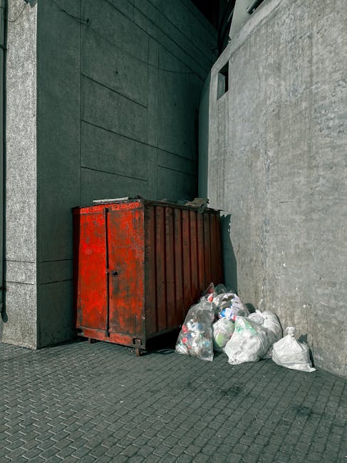 A weathered, red metal skip with visible rust and peeling paint is positioned in a narrow outdoor space between a dark green wall with a smooth finish and a light grey textured concrete wall. The skip is partially filled with miscellaneous waste and is situated on a paved surface made of small, dark grey rectangular bricks. In front of the skip, several transparent plastic bags filled with various types of rubbish, such as paper, plastic bottles, and other debris, are stacked and leaning against its side. The surrounding environment suggests an urban alleyway or service area, possibly used for private waste disposal or an alternative to municipal rubbish collection. Natural light illuminates the scene, highlighting the textures and colors of the skip, waste, and walls, with no other objects or activity visible in the immediate vicinity. The scene reflects typical rubbish removal practices, potentially coordinated by local waste management services or private operators.