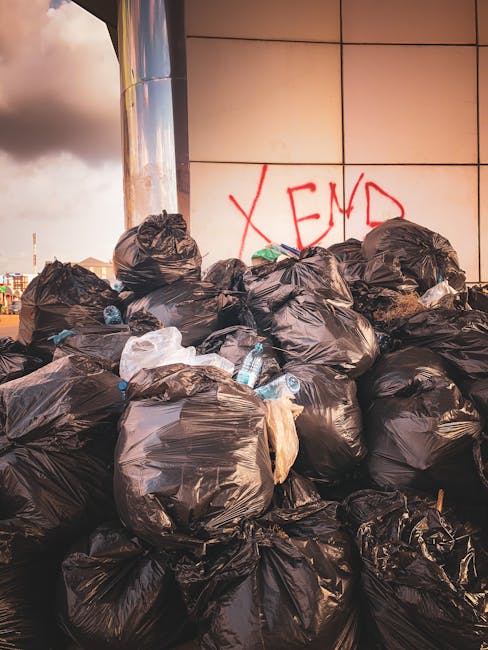 A large pile of black garbage bags, some partially torn or showing contents such as plastic bottles and paper, stacked against a modern building with a reflective, tiled exterior. The bags are placed on a paved surface, with a few scattered loose items visible among them, and the background includes a cloudy sky. On the building's wall, red graffiti spelling 'XEND' is present. This scene illustrates excess waste accumulation often encountered in private rubbish removal or alternative collection contexts, emphasizing the importance of timely rubbish disposal services. The lighting indicates late afternoon or evening, casting a warm hue over the scene, and the overall environment suggests an urban area where waste is temporarily stored pending collection or removal, aligning with themes of independent waste handling and rubbish management.