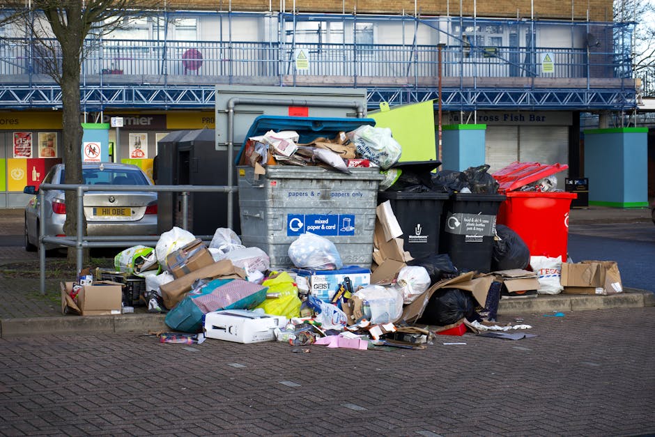 A large pile of black garbage bags, some partially torn or showing contents such as plastic bottles and paper, stacked against a modern building with a reflective, tiled exterior. The bags are placed on a paved surface, with a few scattered loose items visible among them, and the background includes a cloudy sky. On the building's wall, red graffiti spelling 'XEND' is present. This scene illustrates excess waste accumulation often encountered in private rubbish removal or alternative collection contexts, emphasizing the importance of timely rubbish disposal services. The lighting indicates late afternoon or evening, casting a warm hue over the scene, and the overall environment suggests an urban area where waste is temporarily stored pending collection or removal, aligning with themes of independent waste handling and rubbish management.
