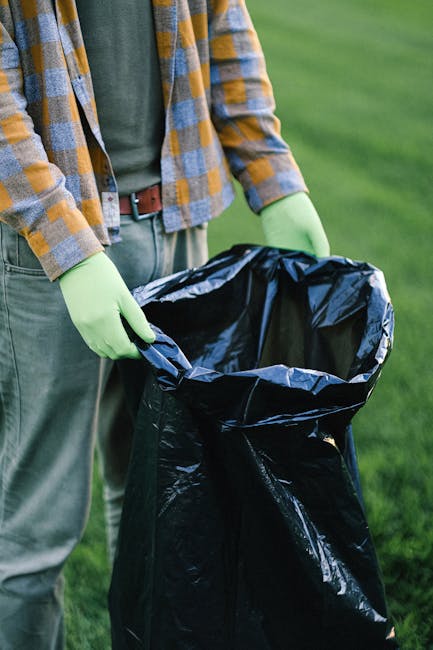 Close-up of a person wearing a plaid shirt, green gloves, and grey trousers, holding open a large black rubbish bag in an outdoor setting with a grassy background. The individual appears to be preparing to dispose of waste, with the bag partially filled and positioned on the ground. The scene suggests an outdoor waste collection or cleaning activity, emphasizing the importance of proper rubbish handling and disposal, consistent with private or independent waste management services like those offered by companies such as rubbishremovalkingstonuponthames.org.uk. The natural daylight and clear environment highlight a routine waste collection task, focusing on the practical use of protective gloves and a sturdy rubbish bag for efficient rubbish removal while supporting overall waste management efforts.