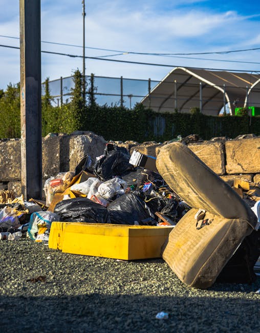 Close-up of a person wearing a plaid shirt, green gloves, and grey trousers, holding open a large black rubbish bag in an outdoor setting with a grassy background. The individual appears to be preparing to dispose of waste, with the bag partially filled and positioned on the ground. The scene suggests an outdoor waste collection or cleaning activity, emphasizing the importance of proper rubbish handling and disposal, consistent with private or independent waste management services like those offered by companies such as rubbishremovalkingstonuponthames.org.uk. The natural daylight and clear environment highlight a routine waste collection task, focusing on the practical use of protective gloves and a sturdy rubbish bag for efficient rubbish removal while supporting overall waste management efforts.