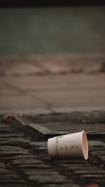 A discarded white paper cup with printed text is lying on its side on a weathered brick pavement, positioned near a drain cover that has parallel metal bars. The background features a section of concrete or stone pavement with a subtle greenish hue, suggesting an outdoor setting such as a street or pathway. The scene is illuminated by natural daylight, highlighting the texture of the bricks and the cup's smooth surface. The placement of the cup and drain cover indicates an area where littering may occur, aligning with waste collection and rubbish removal themes without explicitly suggesting disposal methods.