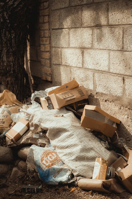 A collection of discarded packaging and waste materials piled on the ground next to a brick wall and a tree trunk. The waste includes several cardboard boxes, some torn and flattened, along with plastic bags and paper wrapping. The materials exhibit various textures and shades of brown and beige, with the cardboard boxes showing printed labels and logos. The scene appears to be outdoors, possibly in a residential or urban area, with some natural light illuminating the waste pile. This image is relevant to rubbish removal services, illustrating the type of waste that might be handled through private or alternative disposal arrangements rather than municipal collection. The waste is loosely stacked without containment, emphasizing the importance of effective rubbish clearance to maintain cleanliness and safety in the environment. The background features a plain, light-colored brick wall, providing a simple and neutral setting that highlights the unmanaged waste in the foreground. The scene supports the concept of rogue or on-site clearance by independent rubbish removal companies, like [COMPANY_NAME], in Kingston upon Thames or similar areas, to assist with unwanted waste disposal outside of standard council services.