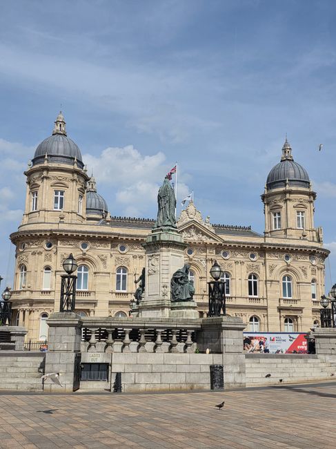A historic sandstone building features ornate architectural details, including rounded turrets topped with domed roofs and decorative stonework. The facade is adorned with multiple arched windows framed in white, and a central entrance flanked by classical columns. In front of the building, there is a stone statue of a seated figure on a pedestal, with an additional standing figure positioned behind. The statue is surrounded by black wrought-iron railings, and three black lampposts are evenly spaced along the edge of the elevated platform. The scene is set on a paved area with a few pigeons, against a backdrop of a bright blue sky with scattered white clouds. The building’s grandeur and the surrounding urban setting suggest a public or civic function, and the area appears clean and well-maintained, ideal for pedestrian movement. This setting exemplifies the type of architecture and outdoor space that often require regular rubbish removal, especially in busy city environments, supported by local services such as those offered by rubbishremovalkingstonuponthames.org.uk.