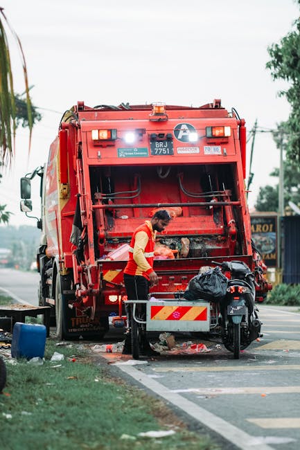 A sanitation worker wearing a high-visibility vest and dark trousers is seen operating a large red refuse truck on the side of a paved urban road. The back of the truck features hydraulic mechanisms and a once-open loading area, with some debris visible on the ground underneath it. The worker appears to be managing waste, with a black plastic bag placed on a nearby metal platform attached to a motorcycle parked adjacent to the truck. In the background, there are trees, utility poles, and a building or fence, suggesting a semi-urban environment. The sky is overcast, casting diffuse lighting over the scene, which emphasizes the metallic surface of the refuse vehicle and the textures of the bags and debris. The scene illustrates a typical waste collection activity, potentially part of an alternative waste handling service aligned with private rubbish removal or on-site clearance, as often employed in local authority or independent rubbish collection operations in Kingston upon Thames.