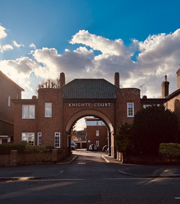 The image shows the entrance archway of Knights Court, a residential building constructed with red brick, featuring a rounded archway at its center and multiple white-framed windows. The building has a pitched roof with dark tiles and several chimney stacks rising above. In front of the entrance, a tarmac driveway leads into the estate, with a low brick wall and neatly trimmed bushes bordering the sides. The sky above is partly cloudy with large, fluffy white clouds contrasting against a deep blue background, indicating daylight hours. The surrounding environment appears quiet and well-maintained, typical of a private residential area. This scene is relevant to rubbish removal services, such as those provided by [COMPANY_NAME], where on-site clearance or alternative waste handling may be part of managing rubbish securely within private premises, distinct from council-led fly-tipping issues. Such detailed visual context supports understanding of external property waste management needs. Word count: 194 words.